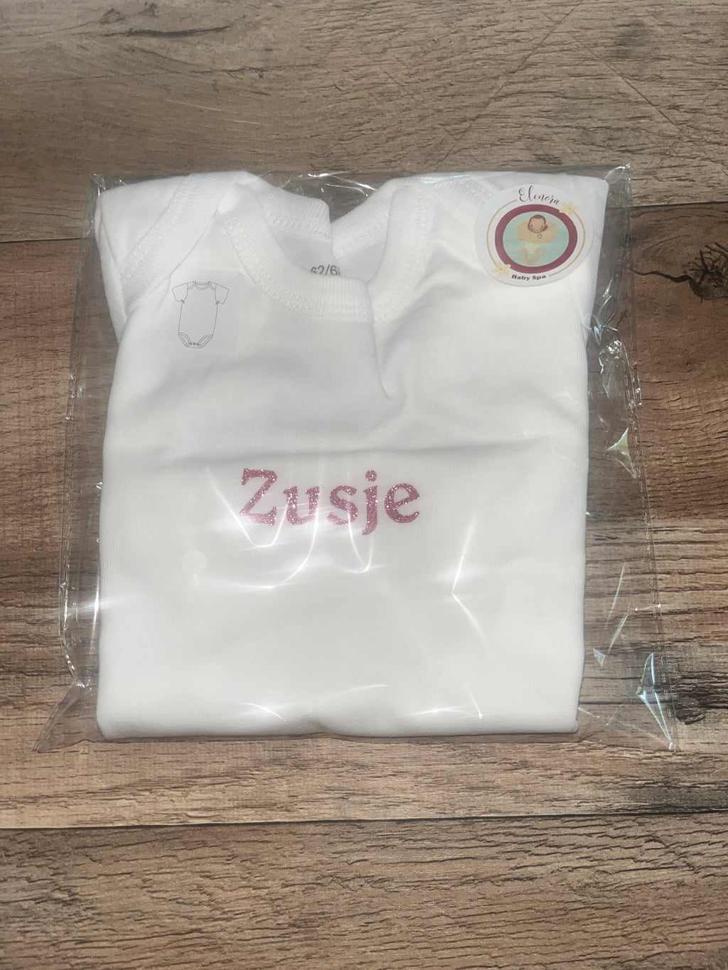 Opgevouwen witte babyromper met roze glittertekst Zusje in doorzichtige verpakking