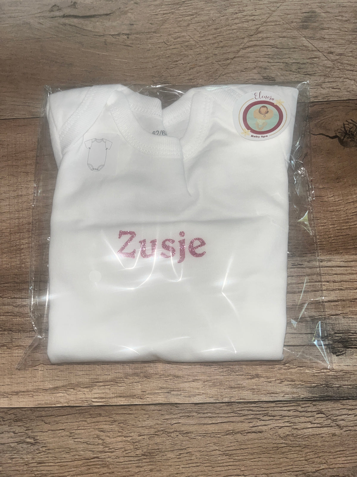 Opgevouwen witte babyromper met roze glittertekst Zusje in doorzichtige verpakking